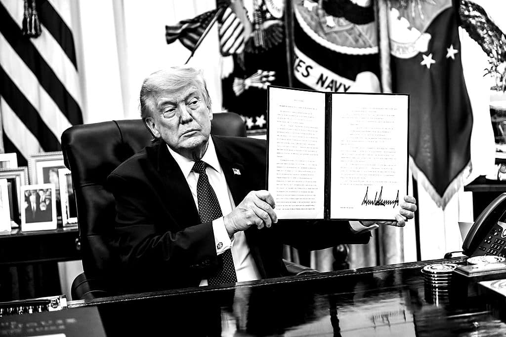 President Trump at the Resolute Desk holding up a signed executive order.